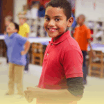 Criança sorridente participando de atividade de Educação Física na sala de aula com outros alunos ao fundo.