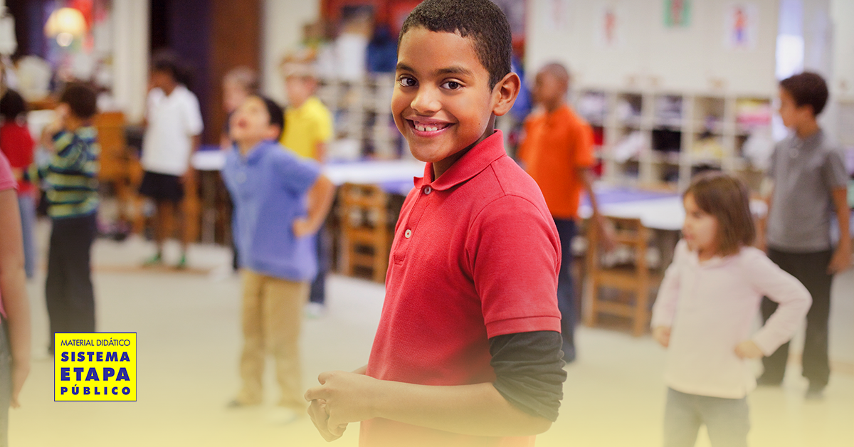 Criança sorridente participando de atividade de Educação Física na sala de aula com outros alunos ao fundo.