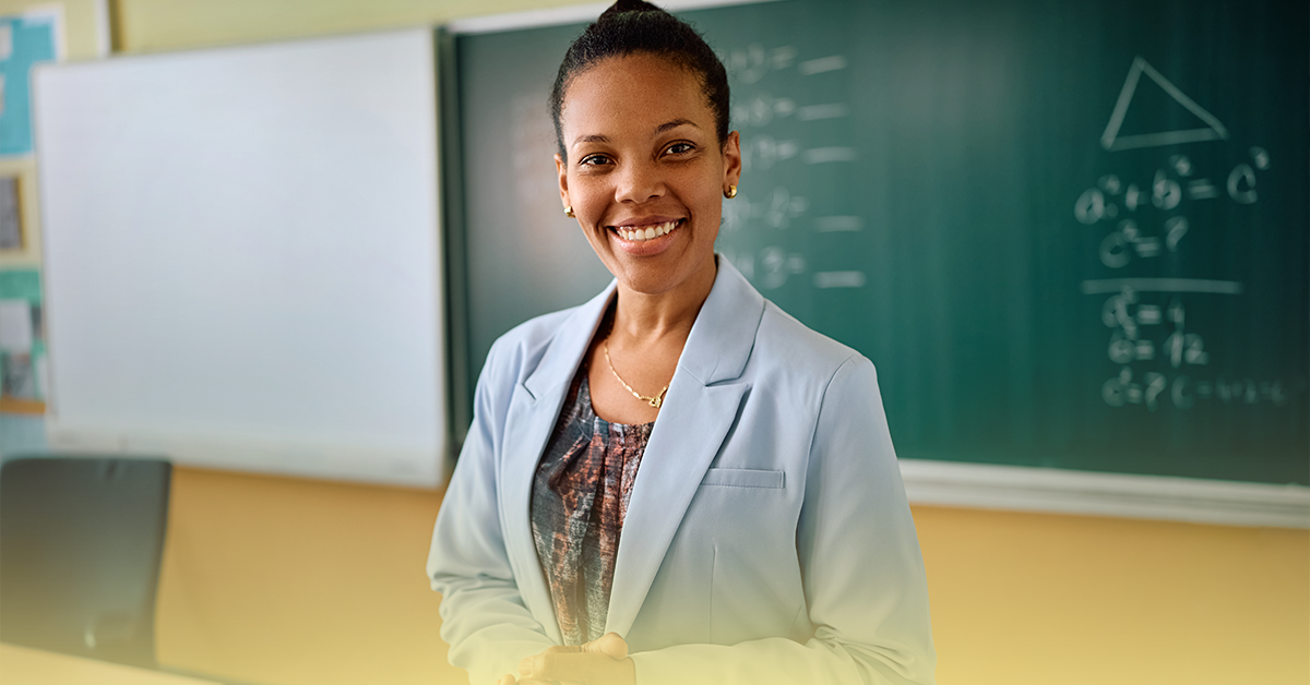 Professora de escola pública sorridente preparando alunos para o SAEB 2025 na sala de aula com quadro-negro ao fundo.
