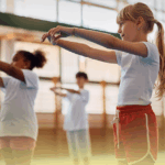 Grupo de crianças praticando Tai Chi Chuan em aula de Educação Física, com movimentos de alongamento e equilíbrio, em ambiente escolar bem iluminado.