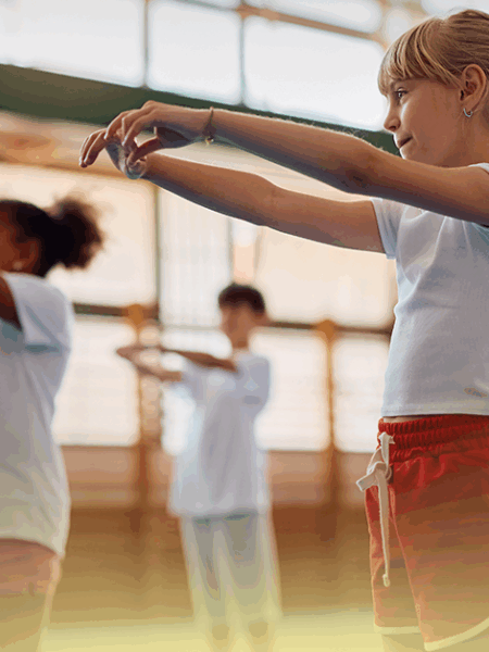 Grupo de crianças praticando Tai Chi Chuan em aula de Educação Física, com movimentos de alongamento e equilíbrio, em ambiente escolar bem iluminado.