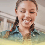Estudante sorrindo enquanto lê um livro em uma biblioteca escolar, representando o incentivo à leitura durante as férias.