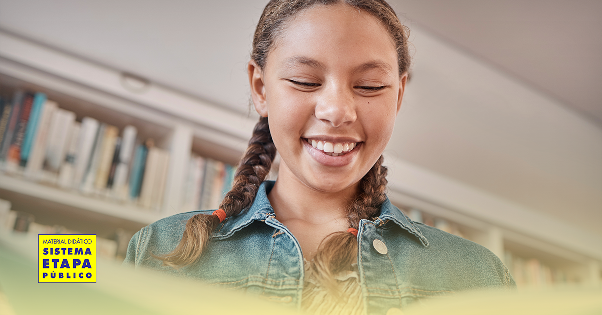 Estudante sorrindo enquanto lê um livro em uma biblioteca escolar, representando o incentivo à leitura durante as férias.