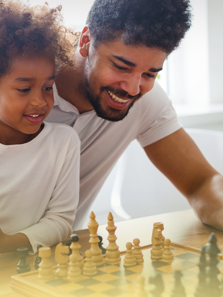 Pai acompanha o filho em atividade lúdica com jogo de tabuleiro, representando experiências cotidianas que estimulam o desenvolvimento cognitivo e socioemocional durante as férias escolares.