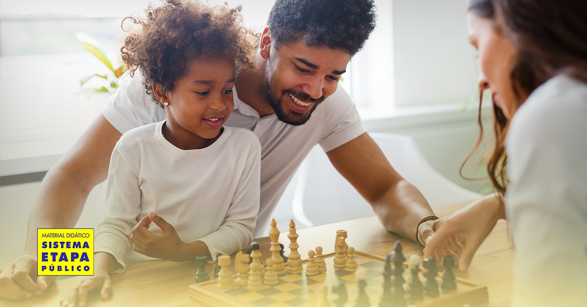Pai acompanha o filho em atividade lúdica com jogo de tabuleiro, representando experiências cotidianas que estimulam o desenvolvimento cognitivo e socioemocional durante as férias escolares.