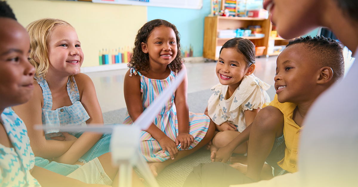 Crianças da Educação Infantil sentadas em roda participam de um momento de conversa e interação, sorrindo e ouvindo a professora em um ambiente escolar acolhedor.