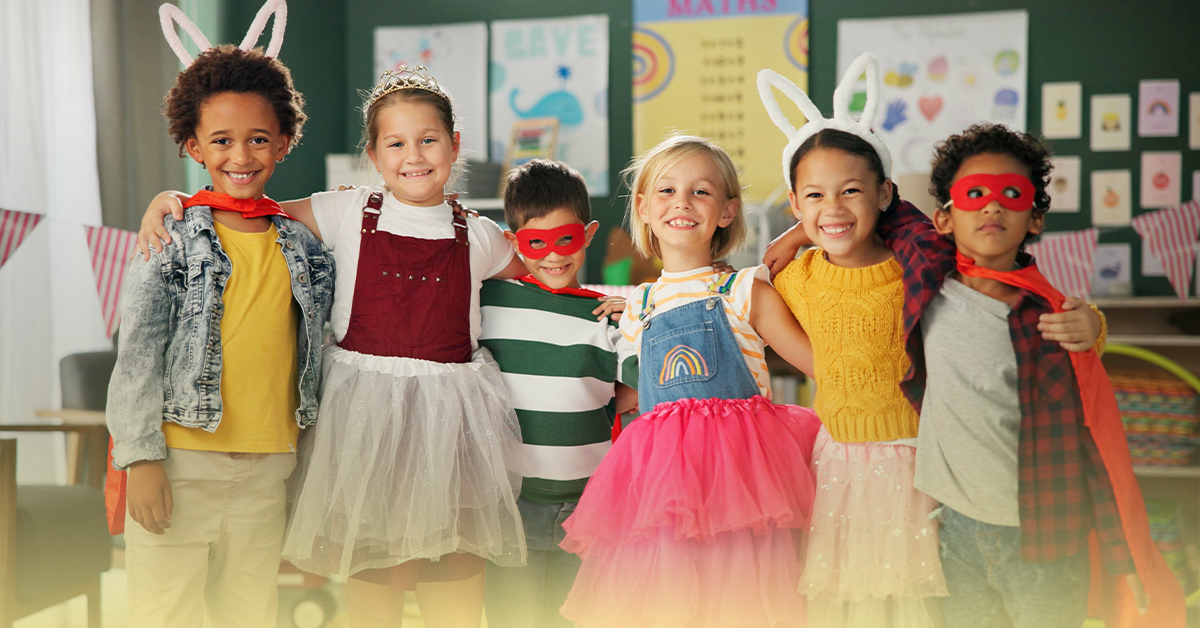 Grupo de crianças fantasiadas sorrindo na sala de aula em uma atividade de carnaval usando orelhas de coelho, tiara e capas de super-herói, com cartazes coloridos ao fundo.