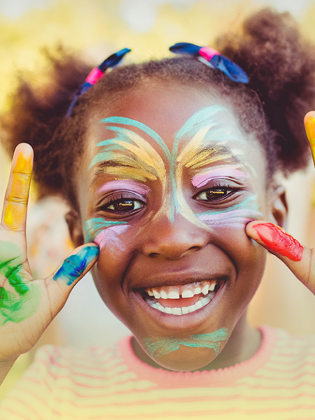 Criança sorrindo com pintura facial colorida em uma atividade de carnaval, mãos abertas com tinta nas palmas e dedos, com outras crianças desfocadas ao fundo.