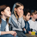 Grupo de crianças assistindo a um filme com expressão de surpresa e curiosidade em um ambiente escuro, como cinema ou sala de projeção.