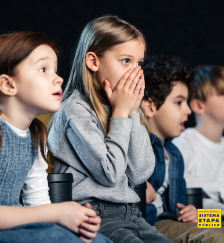 Grupo de crianças assistindo a um filme com expressão de surpresa e curiosidade em um ambiente escuro, como cinema ou sala de projeção.