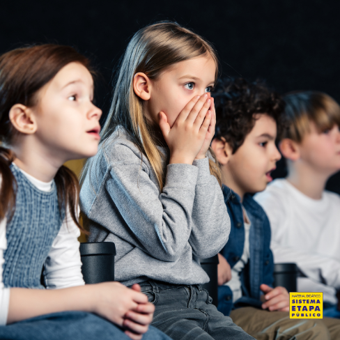 Grupo de crianças assistindo a um filme com expressão de surpresa e curiosidade em um ambiente escuro, como cinema ou sala de projeção.