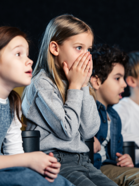 Grupo de crianças assistindo a um filme com expressão de surpresa e curiosidade em um ambiente escuro, como cinema ou sala de projeção.
